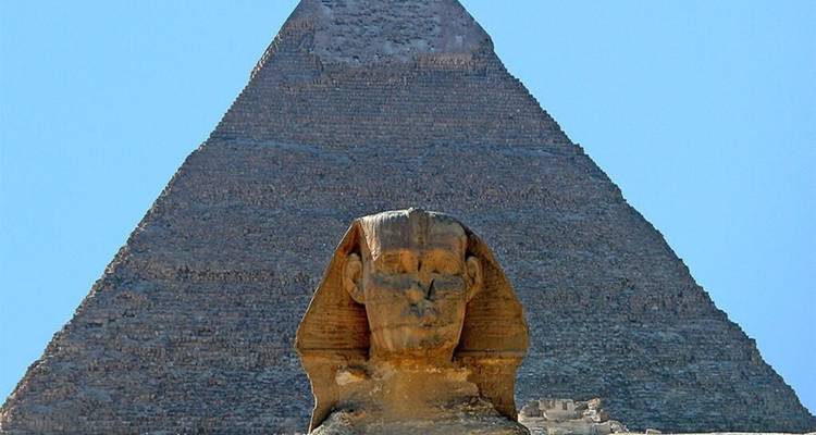 Close-up van de Grote Sfinx met een kolossale piramide die erachter omhoog rijst tegen een heldere blauwe hemel.