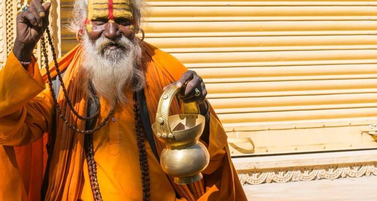 Portrait of a colorfully dressed Sadhu holding prayer beads and a brass vessel.