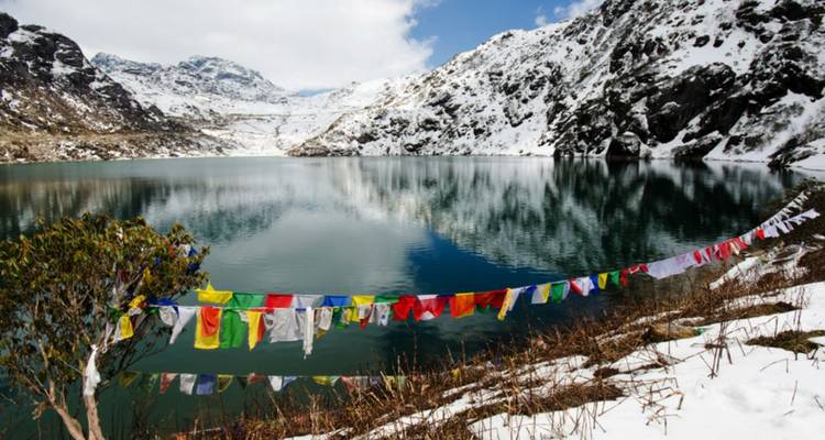Mirror-still alpine lake ringed by snowy slopes, reflecting mountains and prayer flags fluttering in the breeze