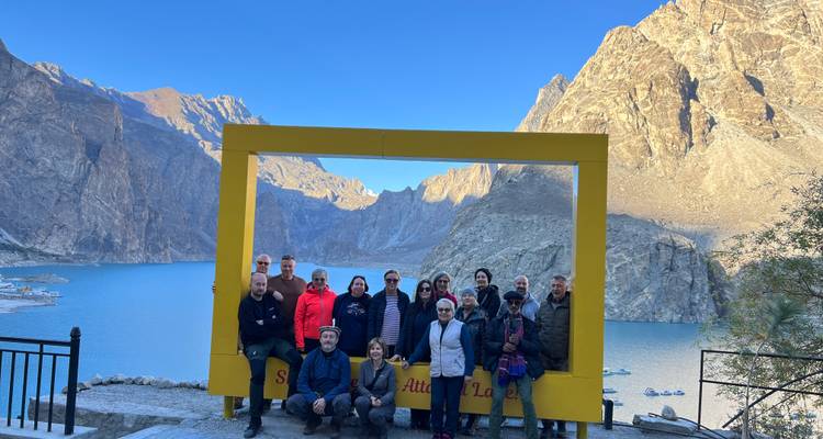 Un groupe de touristes posant à l'intérieur d'une grande structure de cadre photographique jaune dominant le lac Attabad d'un bleu éclatant et les montagnes rocheuses environnantes.