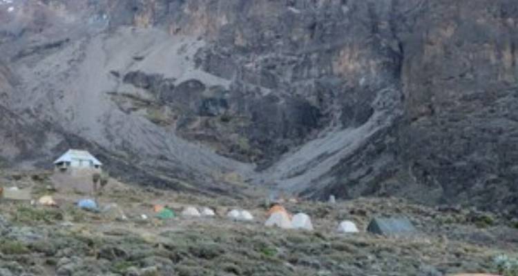 Vue lointaine d'un campement rocheux gris avec des tentes dispersées au pied d'une paroi volcanique escarpée