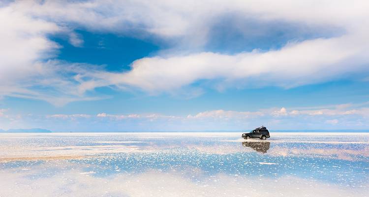 4x4-Fahrzeug, das über einen spiegelglatten Salzsee fährt und einen weiten blauen Himmel mit zarten Wolken reflektiert.