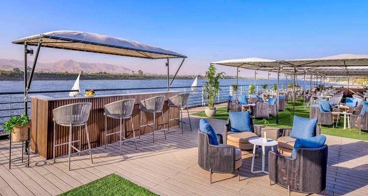 Stilvolle Open-Air-Bar und Lounge-Deck auf einem Nilkreuzfahrtschiff mit Fluss und Bergen im Hintergrund.