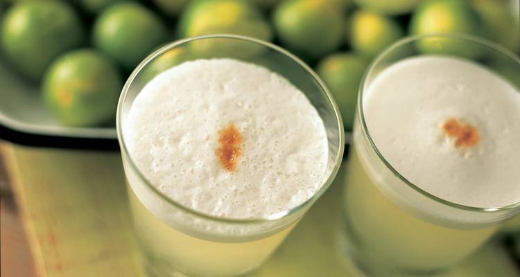 Two frothy pisco sours in clear glasses with limes blurred in the background on a wooden surface.