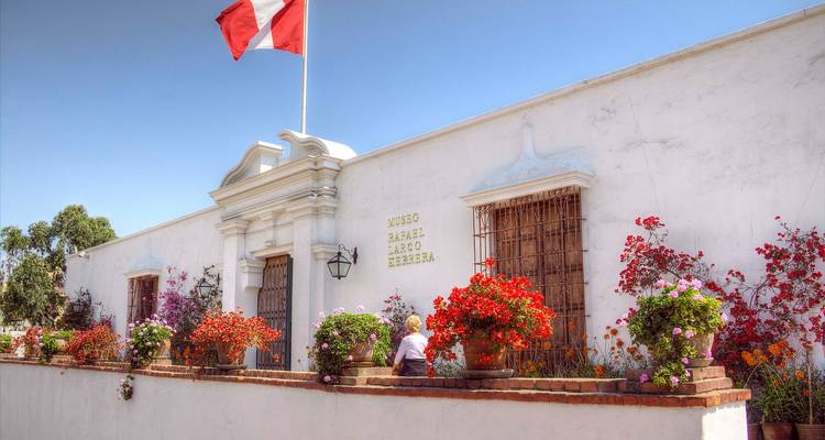 White colonial-era Larco Museum façade in Lima framed by colourful flowerpots and a Peruvian flag waving against a blue sky.