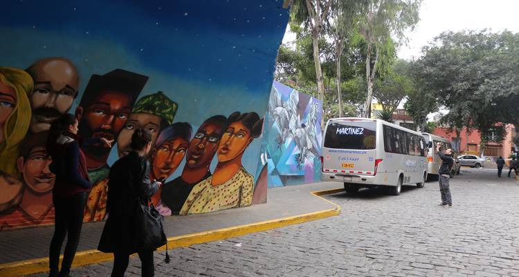 Tourists photograph vibrant street murals beneath an urban overpass while a small bus turns onto a cobblestone street.