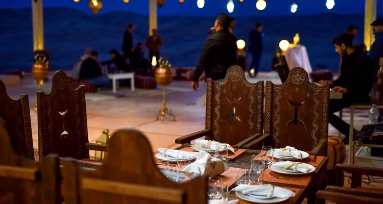 Vista cercana de mesas de comedor elegantemente preparadas mientras los huéspedes se mezclan en una carpa abierta iluminada por linternas por la noche.