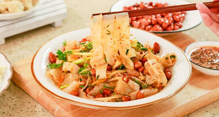 Bowl of spicy Liangpi cold noodles garnished with peanuts and herbs being lifted with chopsticks.