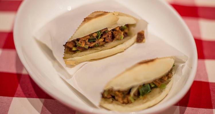 Sliced Roujiamo meat bun rests on a white dish lined with parchment on a checkered cloth.