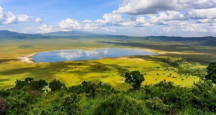 Vue panoramique sur le plancher verdoyant et le lac central du Cratère du Ngorongoro sous des nuages épars.