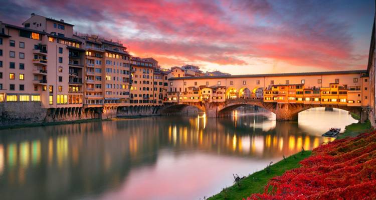 Ponte Vecchio weerspiegeld in de Arno onder een levendige zonsonderganghemel getint met rode en paarse tinten.