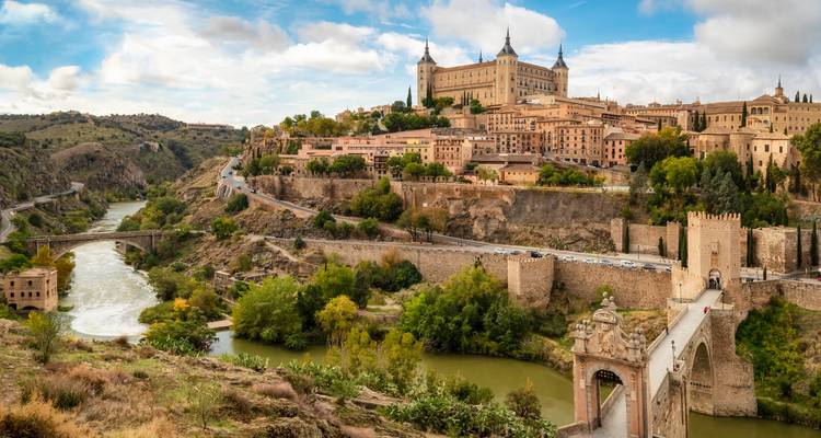 Panoramablick auf eine historische spanische Bergstadt mit einem imposanten Festungspalast, der einen gewundenen Fluss und eine alte Steinbrücke überblickt, eingerahmt von grünen Hügeln unter einem teilweise bewölkten Himmel.