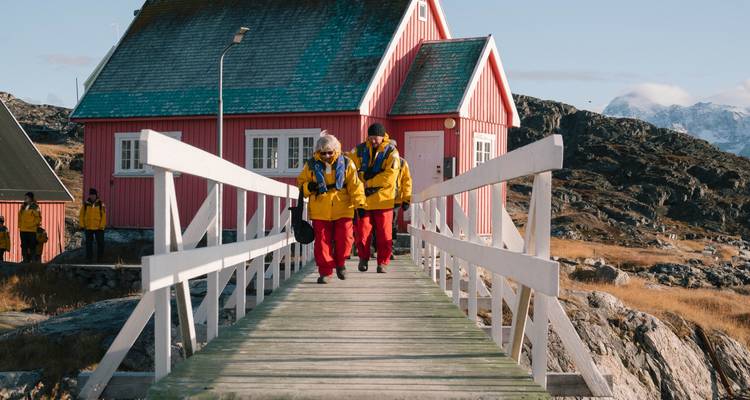 Expeditionsgäste in leuchtend farbigen Parkas gehen über eine hölzerne Brücke zu einer roten arktischen Hütte.