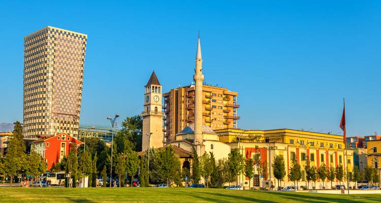 Le skyline de Tirana présentant la Mosquée Et'hem Bey, la tour de l'horloge et les bâtiments modernes sous un ciel bleu dégagé.