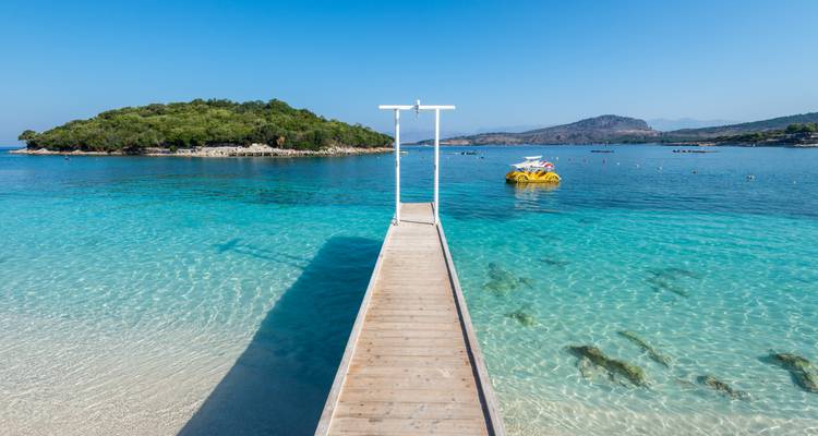 Eau d'un bleu cristallin et une jetée en bois menant vers un petit îlot boisé à Ksamil.