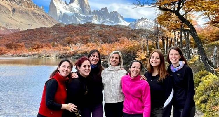 Group of female hikers smile beside a lake with Mount Fitz Roy towering behind in autumn colors.