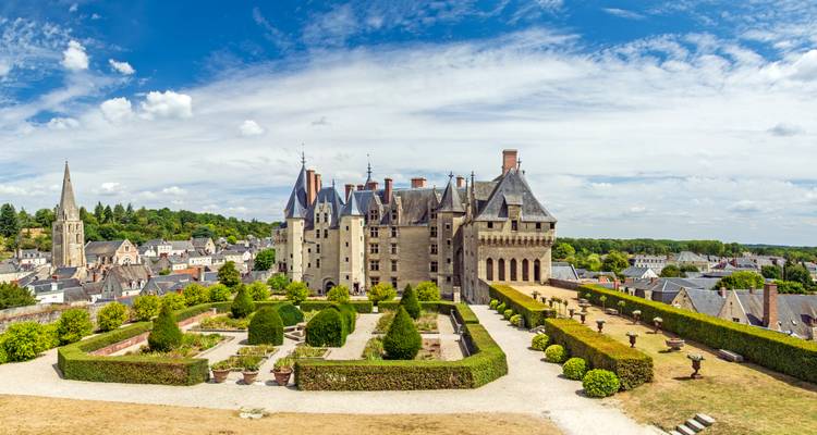 A well-preserved stone château dominates a manicured garden and village beneath a bright blue sky.