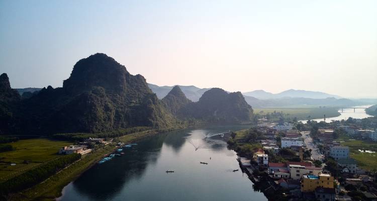 Ein ruhiger Fluss windet sich an Karstbergen und einem kleinen vietnamesischen Dorf unter einem dunstigen Himmel vorbei.