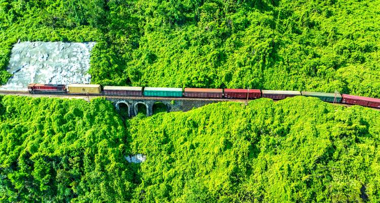 Ein farbenfroher Güterzug überquert ein steinernes Viadukt durch dichten, grünen Dschungel aus der Vogelperspektive.