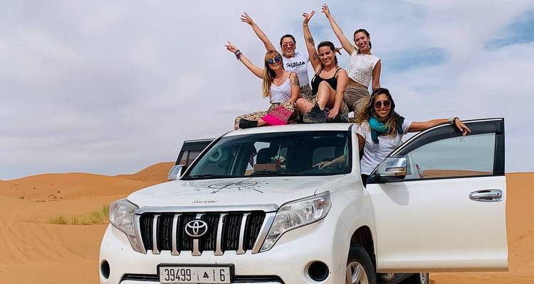 Lächelnde Freunde posieren auf einem weißen 4x4 inmitten von sanften Sanddünen der Sahara.