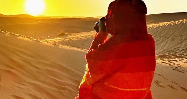 Eine in einen roten Schal gehüllte Gestalt fotografiert die Sonne, die über goldenen Sahara-Dünen aufgeht.