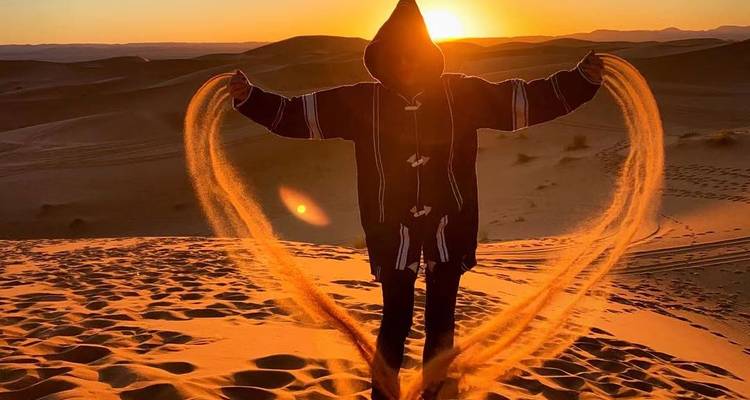 Person in hooded cloak tossing arcs of glowing sand into the air at sunrise on the dunes.