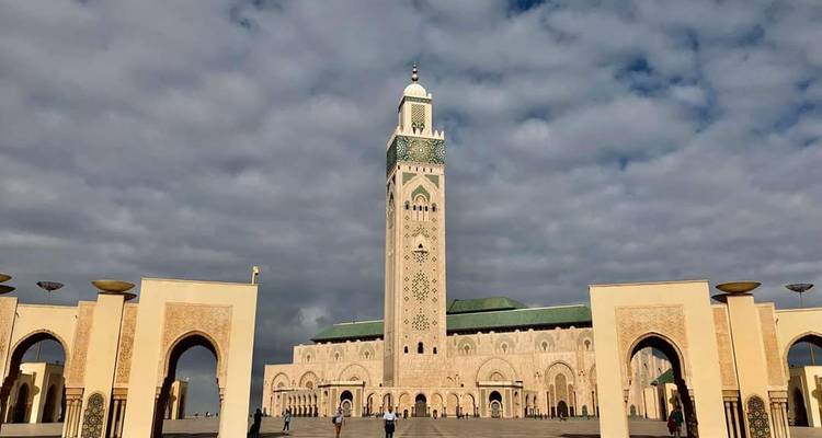 Weiter Platzblick der Hassan-II-Moschee mit kunstvollen Bögen unter einem dramatischen, wolkenbedeckten Himmel.