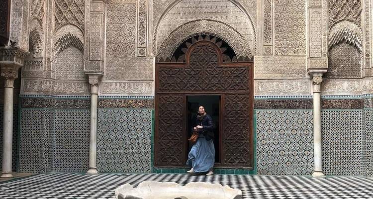 Frau, die durch einen kunstvoll gefliesten Eingang in einer historischen Madrasa mit gemusterten Wänden und Brunnen austritt.