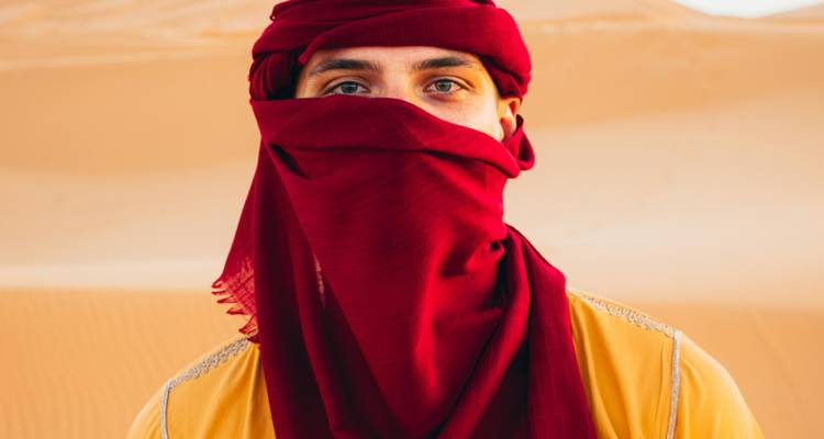 Porträt eines Mannes, der einen dunkelroten Turban und einen Gesichtsschleier trägt und vor weichen Dünen der Wüste steht.