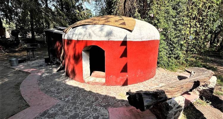 Kleine rot-weiße Schwitzhütte mit Kuppeldach, umgeben von Bäumen mit Steinpfad.