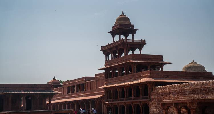 Historischer roter Backsteinturm und Säulenhallen von Fatehpur Sikri vor einem blassen blauen Himmel.