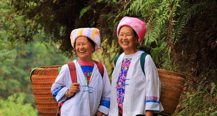 Deux femmes souriantes issues de minorités vêtues de costumes traditionnels portant des paniers tissés dans une végétation luxuriante
