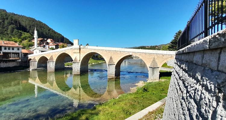 Historic stone arch bridge spanning a clear river with reflections, set against forested hills and bright blue sky.