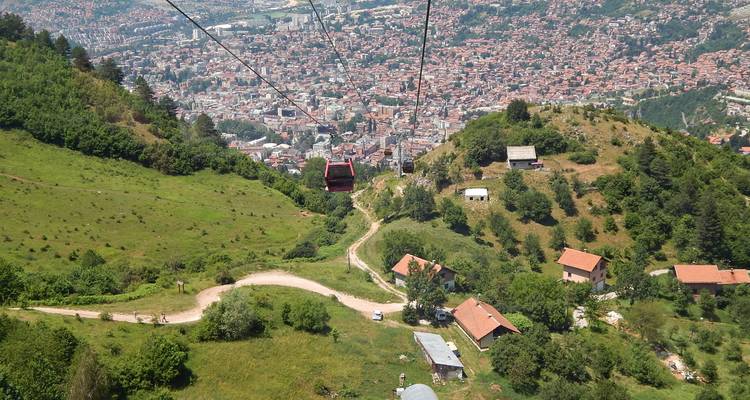 Cable car descending a green hillside toward a sprawling cityscape nestled in a valley.