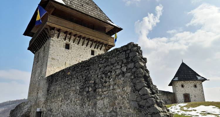 Stone fortress tower flying Bosnian flags under a partly cloudy sky with patches of snow on the ground.
