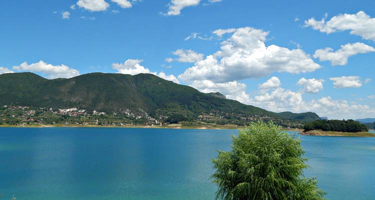Vibrant turquoise lake bordered by forested hills and distant mountains under blue sky