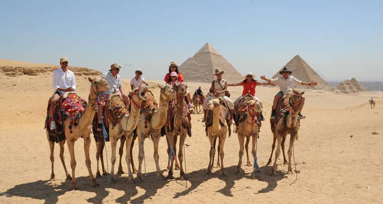Sept cavaliers de chameaux s'alignent avec trois pyramides s'élevant derrière eux dans le désert.