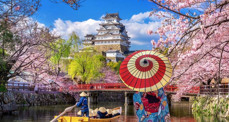 Scène encadrée par les fleurs de cerisier d'un bateau avec des passagers portant des chapeaux de paille et une figure vêtue d'un kimono sous un parasol, avec le château de Himeji s'élevant derrière un pont rouge.