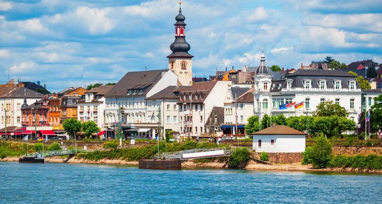 Charmante petite ville au bord du Rhin avec des maisons aux couleurs pastel, une église surmontée d'un dôme en oignon noir, et des collines verdoyantes en arrière-plan, vue de l'autre côté du fleuve par une belle journée.