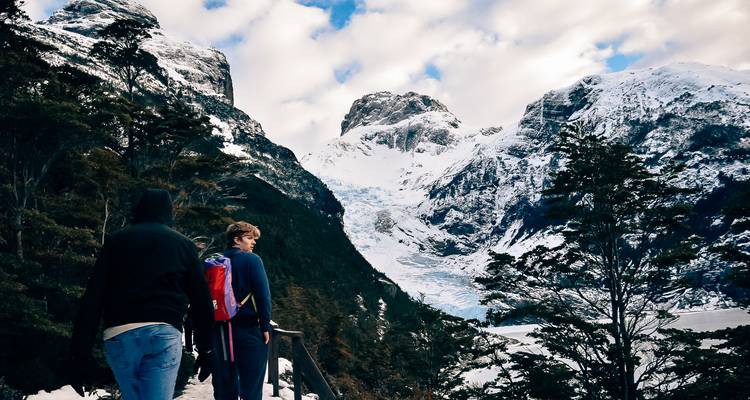 Sendero del valle cubierto de nieve con dos trekkers acercándose a un glaciar colgante bajo cielos parcialmente nublados.