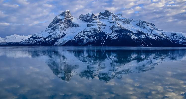 Cresta montañosa cubierta de nieve perfectamente reflejada en un fiordo tranquilo como un espejo al atardecer con nubes suaves arriba.