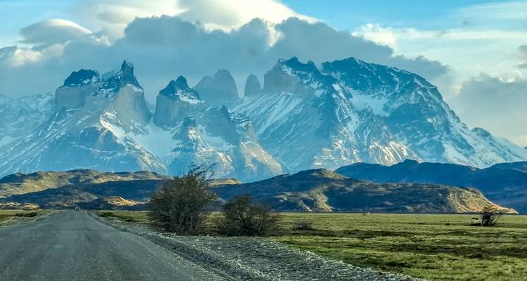 Largo camino de grava recto que se dirige hacia los picos nevados de las Torres del Paine con llanuras verdes a ambos lados.
