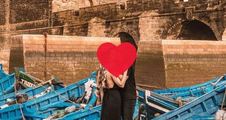 Couple s'embrassant à côté de bateaux de pêche bleus et de remparts en pierre avec un graphique en forme de cœur masquant les visages.
