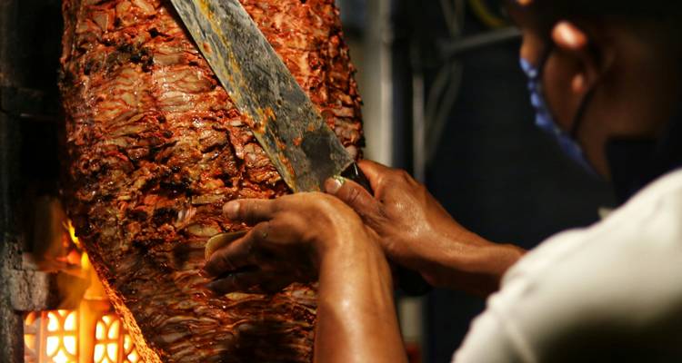 Der Koch schneidet einen vertikalen Spieß mit mariniertem Schweinefleisch "al pastor" neben glühenden Flammen auf.