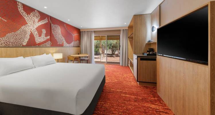 Chambre d'hôtel moderne avec un grand lit blanc, un tapis à motifs rouges et un mur de caractéristique d'art autochtone, donnant sur un jardin désertique par des portes coulissantes en verre.