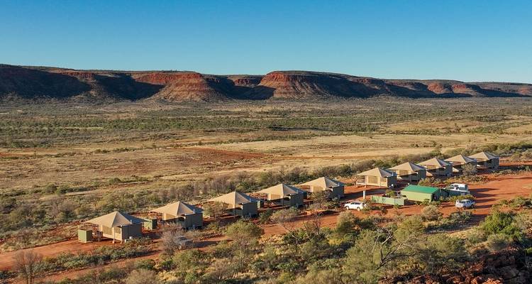 Une rangée de petits éco-chalets en toile repose sur un sol désertique rougeâtre sous des mesas aux sommets plats et un ciel bleu dégagé.