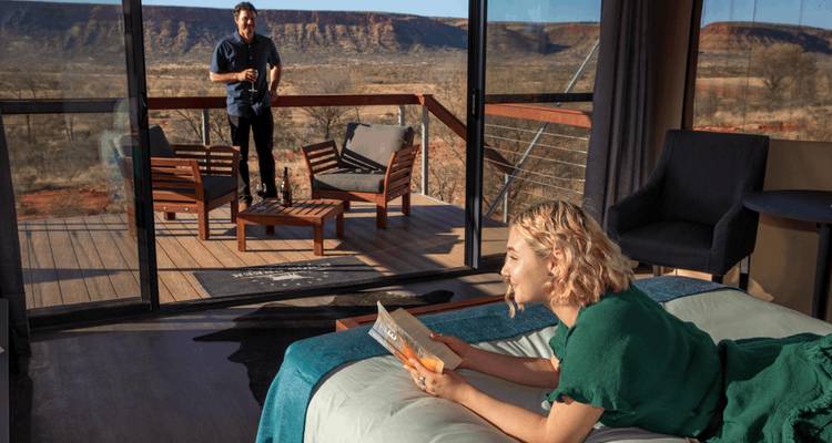 À l'intérieur d'une tente de luxe, une femme lit sur le lit tandis qu'un homme se tient sur la terrasse surplombant les falaises du désert.