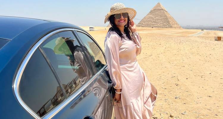 Femme élégante vêtue d'une robe fluide se tenant à côté d'une automobile avec une vue lointaine de pyramide.