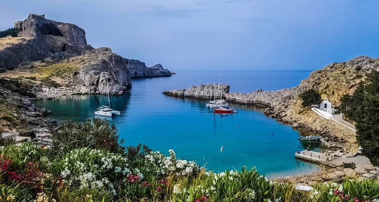 Idyllische Bucht mit klarem blauem Wasser, Yachten, einer kleinen weißen Kapelle und Wildblumen, die die Szenerie überblicken
