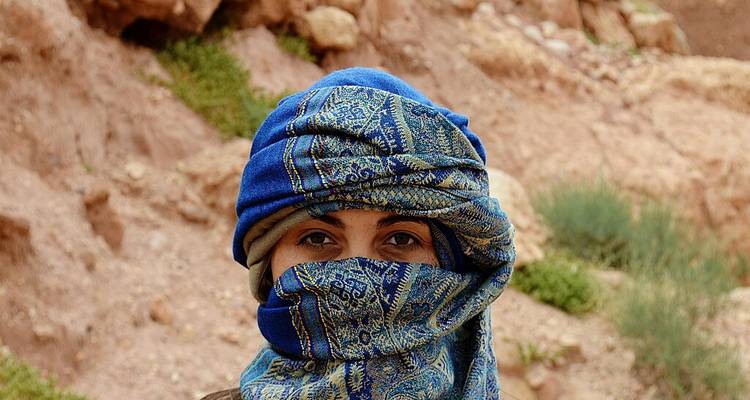 Portrait rapproché d'un voyageur portant un turban bleu à motifs sur un fond de désert rocheux.